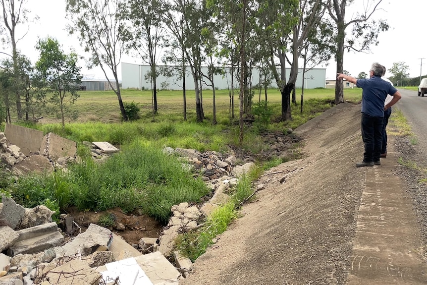 Residents stand on the edge of a causeway in front of infrastructure damaged by flooding.