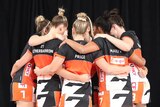 The Giants netball team stand in a huddle with their arms around each other. They are wearing their orange and grey uniform.