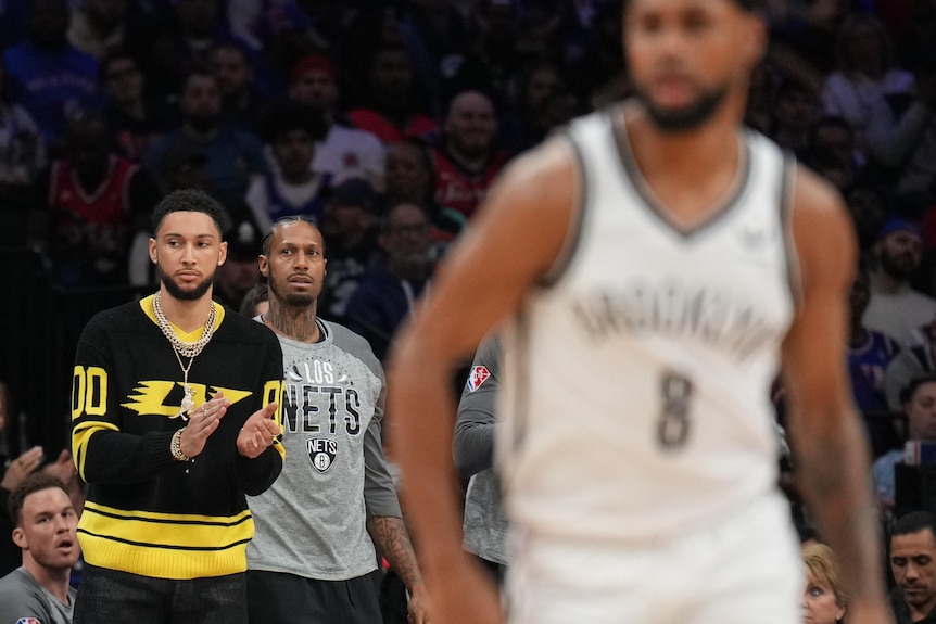 NBA player Ben Simmons stands on the sidelines, clapping. Brooklyn Nets teammate Patty Mills is blurry in the foreground.