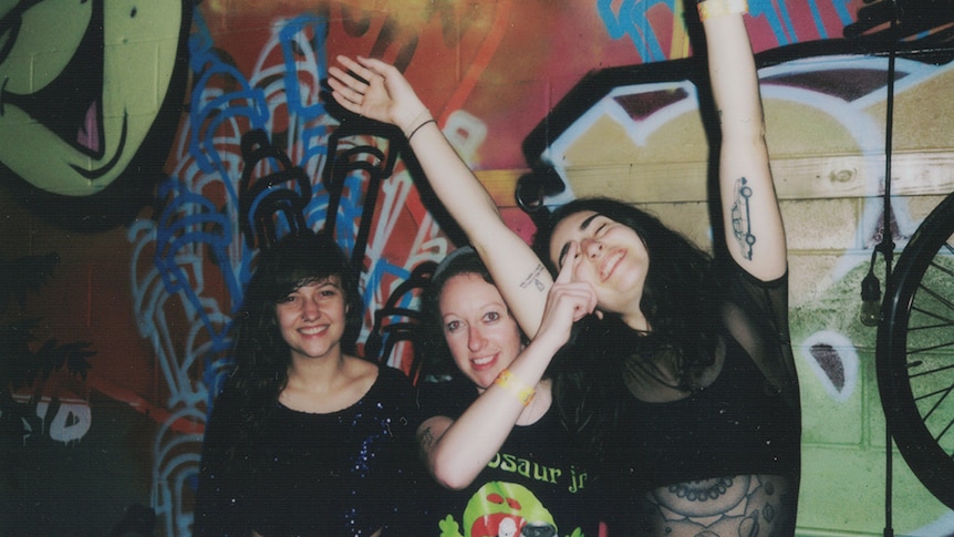 A 2017 polaroid shot of Melbourne band Camp Cope posing against a graffiti'd wall
