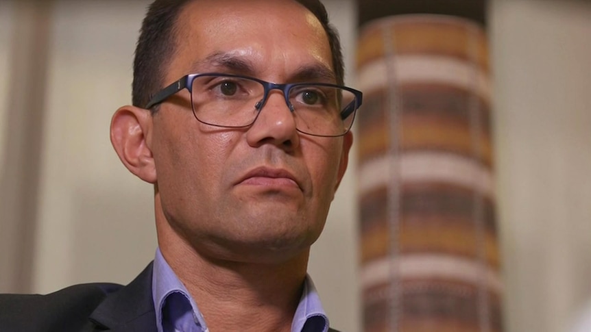 An Aboriginal  man wearing glasses standing in front of a totem