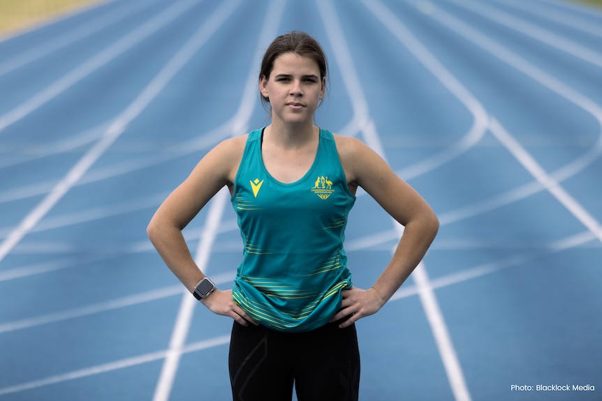 Indi is standing with her hands on her hips on the running tracks. She is wearing a teal blue/green Commonwealth jersey