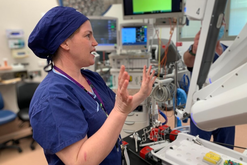 A woman in scrubs and a scrub hat talks with hands up standing in theatre with a surgery robot