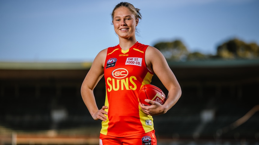 A Gold Coast Suns AFLW recruit stands with the ball held in her left hand.