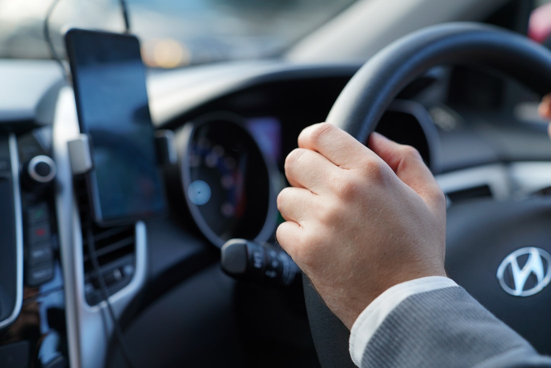 A close-up on a man's hand gripping the steering wheel. A mobile phone sits in its mount on the dashboard.
