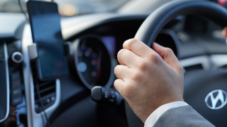 A close-up on a man's hand gripping the steering wheel. A mobile phone sits in its mount on the dashboard.