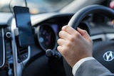 A close-up on a man's hand gripping the steering wheel. A mobile phone sits in its mount on the dashboard.
