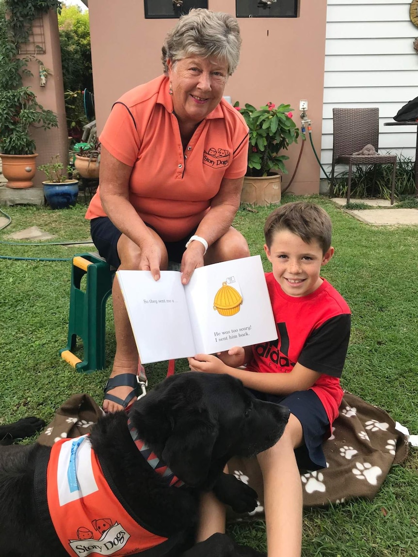 A woman and a child hold a book up next to a sitting dog.