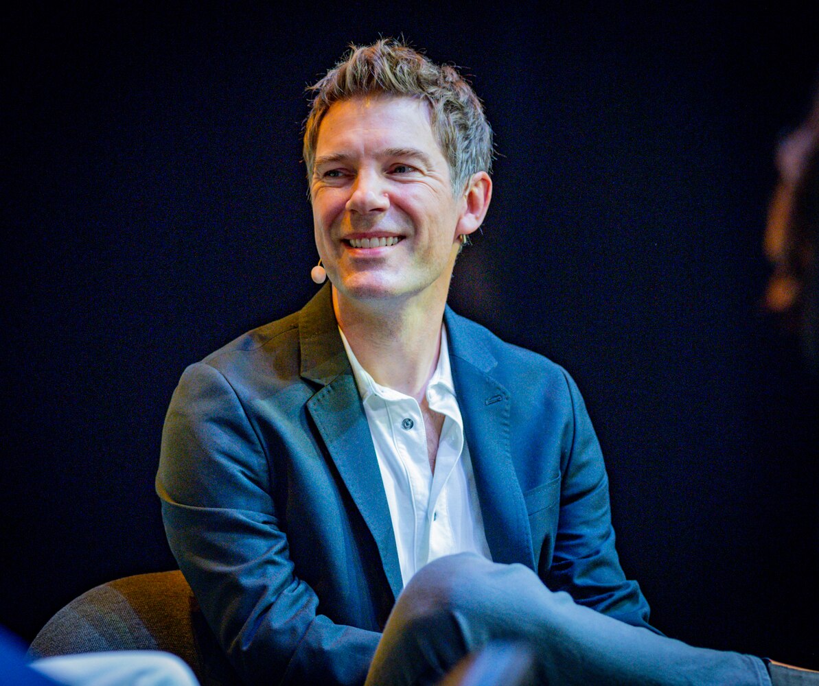 Per Saari on stage smiling wearing a lapel mic and suit jacket and shirt with one leg crossed over the other