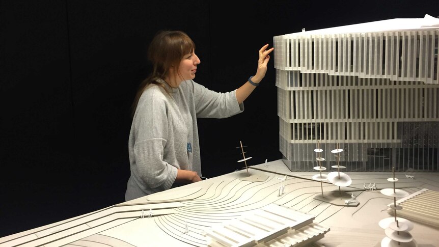 A brown-haired woman stands next to an architect's model of a library, raising her hand to touch its wall.