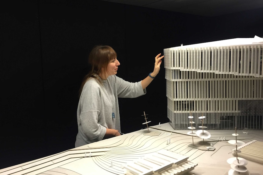 A brown-haired woman stands next to an architect's model of a library, raising her hand to touch its wall.