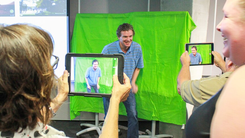 A man stands in front of a piece of green cloth while people with tablets and iPads take his photo