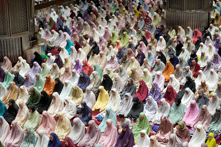 A large group of women face the same way as they pray inside a mosque while wearing colouful clothes and headscarves.