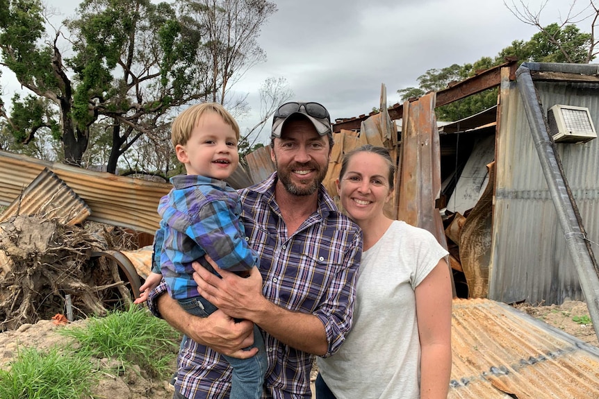 Dan Tarasenko and his partner and son and stand smiling in front of a shed that's been fire-damaged.