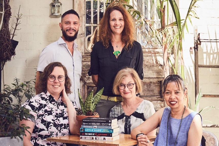 The judges sit around an outdoor coffee table in a leafy sunlit courtyard.