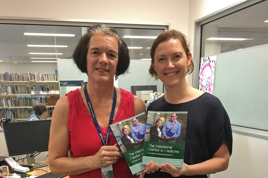 Two women stand in front of a library holding copies of a book about mentoring for medical professionals