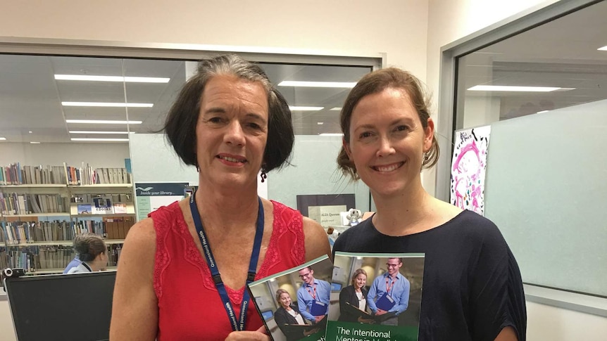 Two women stand in front of a library holding copies of a book about mentoring for medical professionals