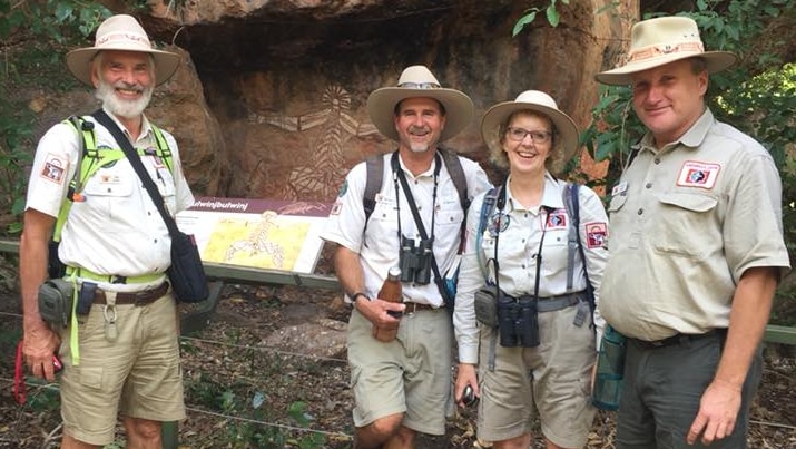 Four people in hats stand in front of Aboriginal rock art