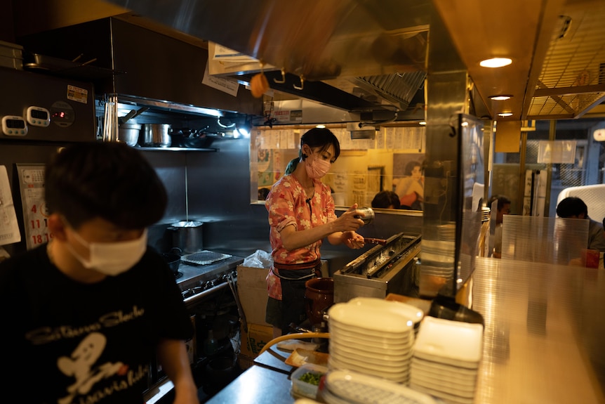 A masked woman grilling meat in a commercial kitchen 