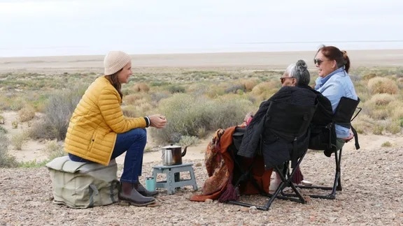 Three women in winter clothing sit on portable chairs and chat in a desert landscape.