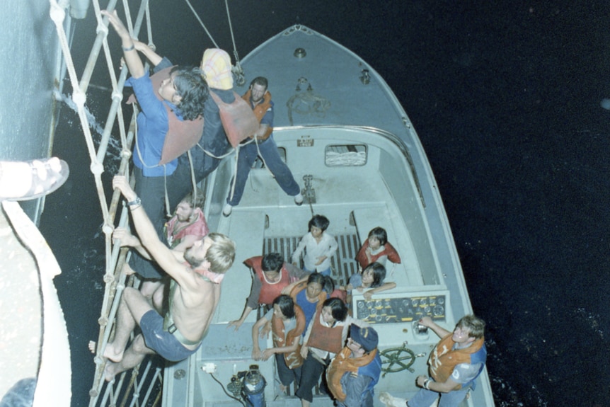 A group of Vietnamese refugees in a tender boat and wearing life vests start to climb a later up to a the deck of a warship