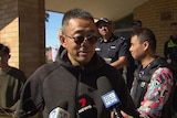 A man wearing a black stop and sunglasses speaks into media microphones.
