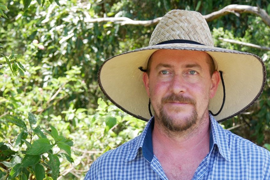 A man standing in bushland who has a beard and is wearing a wide brimmed hat