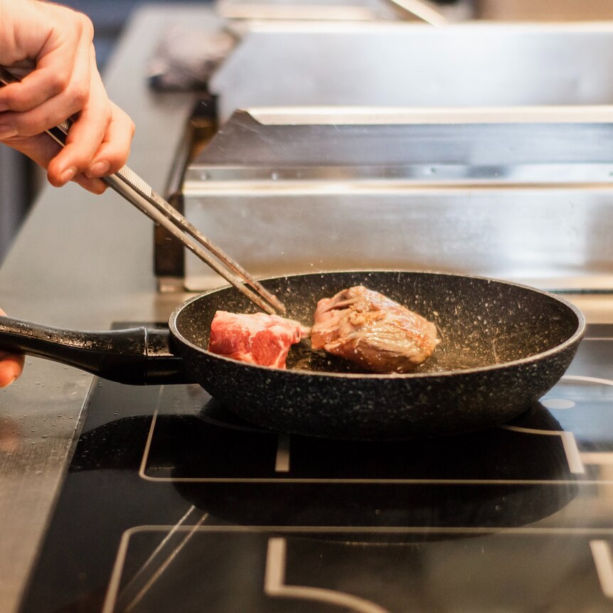 A man uses chopsticks to rotate a hunk of meat in a frypan on an induction stovetop.