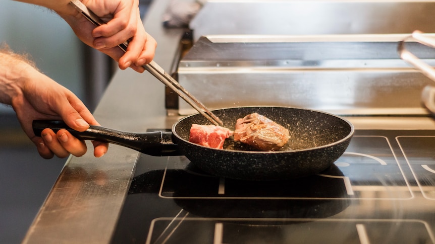 A man uses chopsticks to rotate a hunk of meat in a frypan on an induction stovetop.