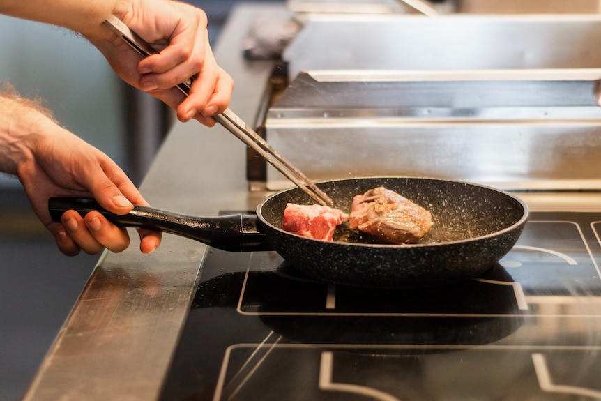 Мужчина палочками для еды переворачивает кусок мяса в сковороде на индукционной плите.