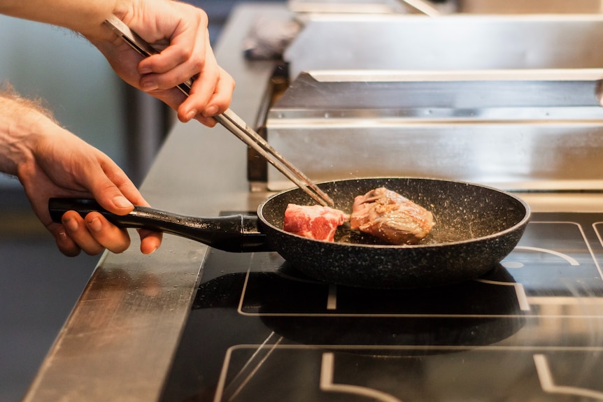 A man uses chopsticks to rotate a hunk of meat in a frypan on an induction stovetop.