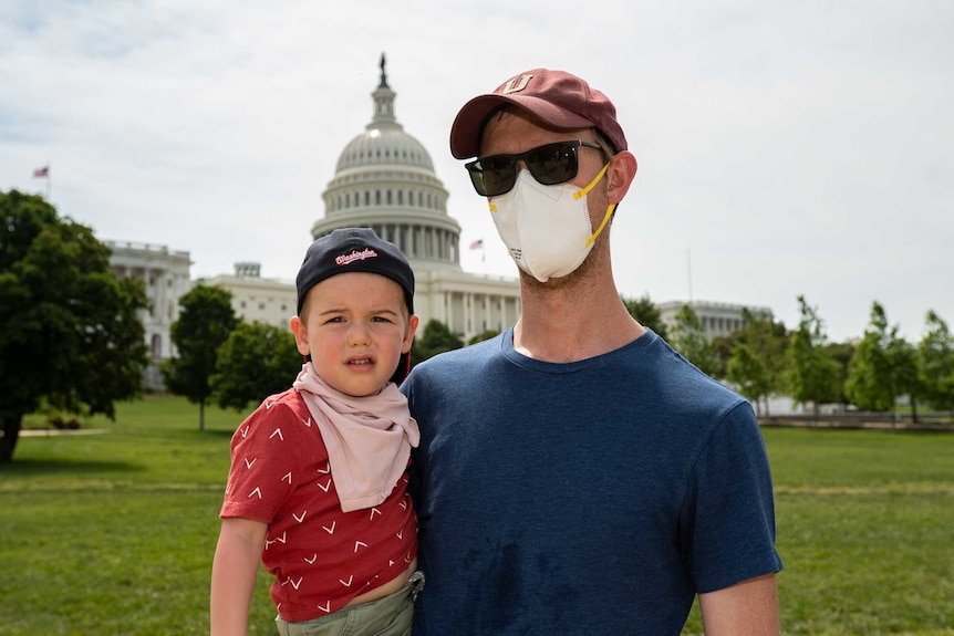 A man in a face mask stands on the lawn outside the capitol building holding his young son