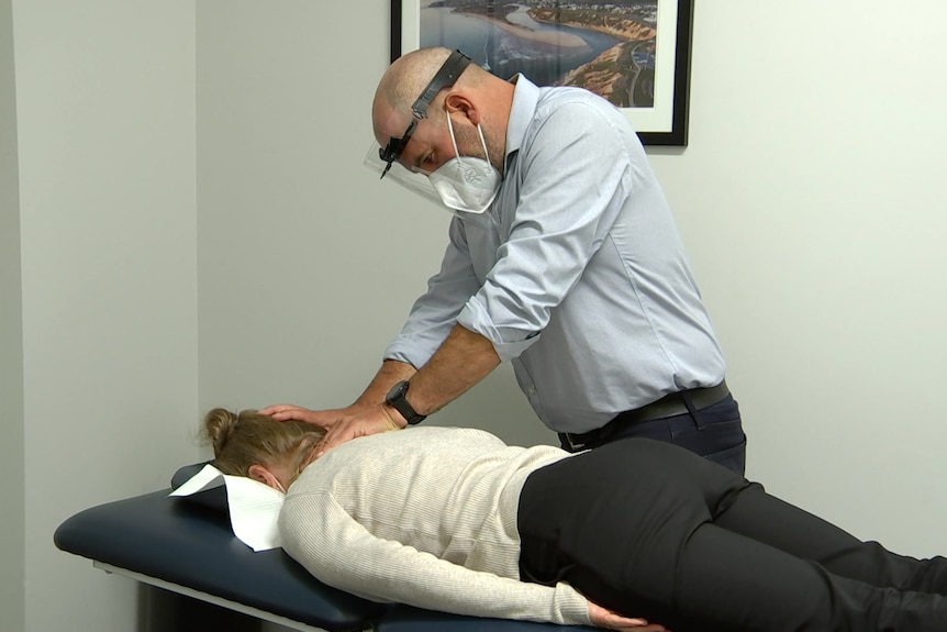 A man wearing a face shield and face mask while touching a patient's neck on a massage table