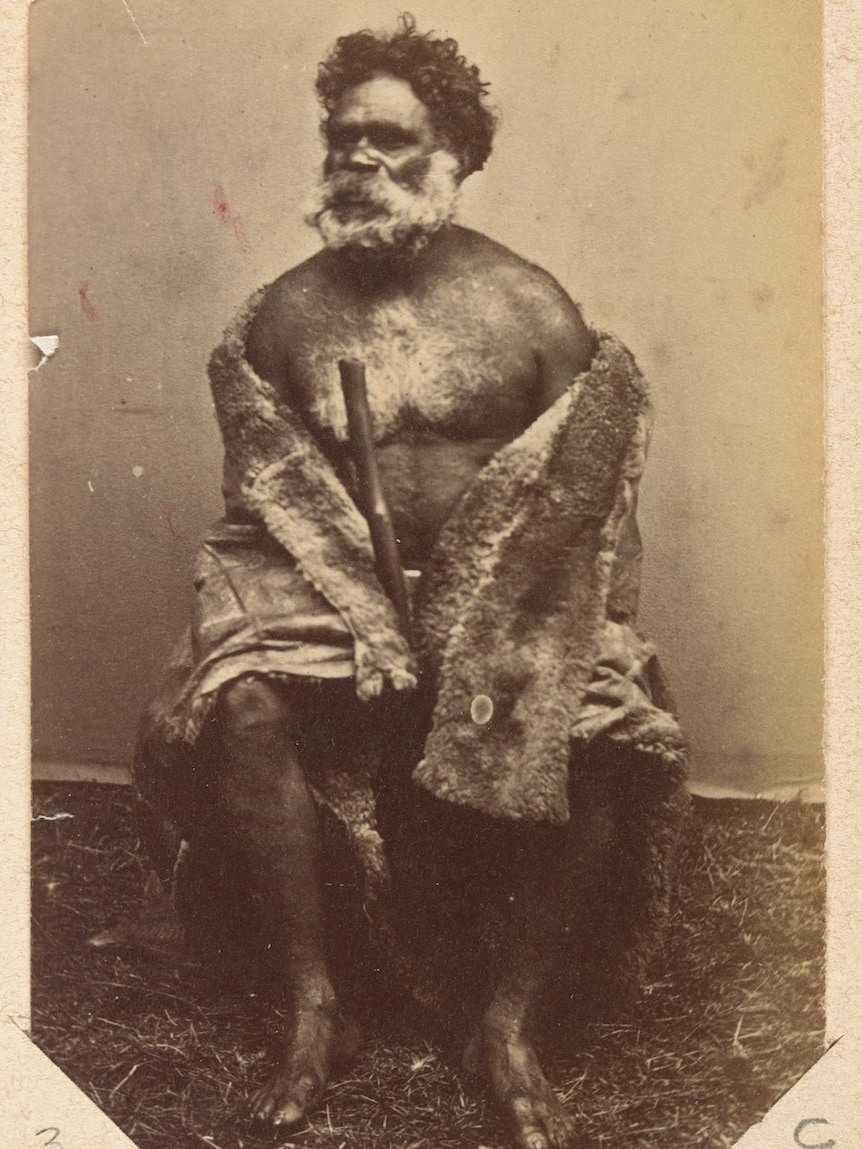 Indigenous man with full grey beard looks into the distance. He sits bare-footed, wrapped in animal fur and holding large stick