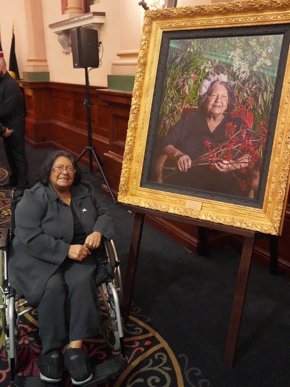 An older woman in a wheelchair sits beside her photographic portrait, which is gold-framed.