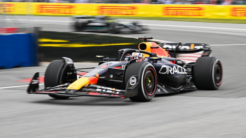 Max Verstappen in his navy blue F1 car, turns through a corner as he races, with one car chasing behind.