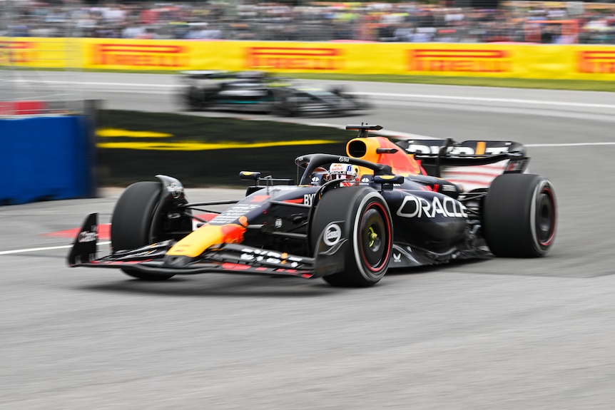 Max Verstappen in his navy blue F1 car, turns through a corner as he races, with one car chasing behind.