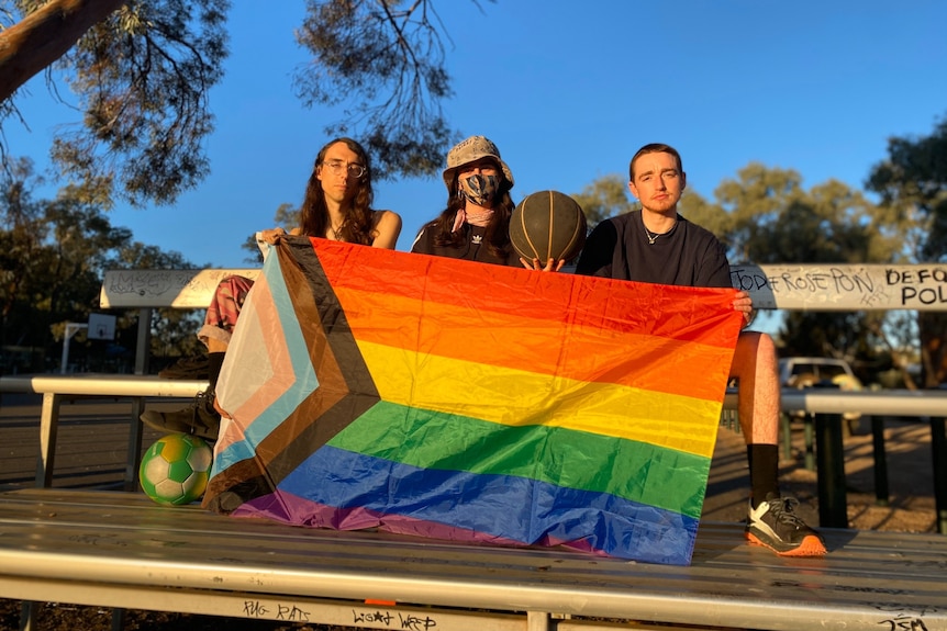 three people sitting on silver benches at a basketball court, holding a large rainbow flag