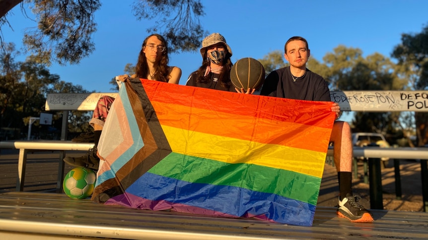 three people sitting on silver benches at a basketball court, holding a large rainbow flag