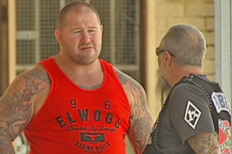 A man in a red singlet stands talking to another man who is wearing a bikie jacket.