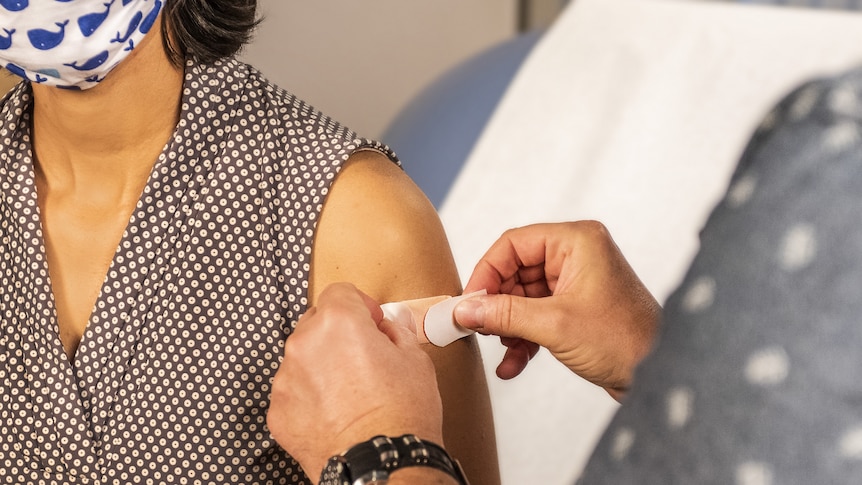 A person applying a bandage on the shoulder of another person who is wearing a mask