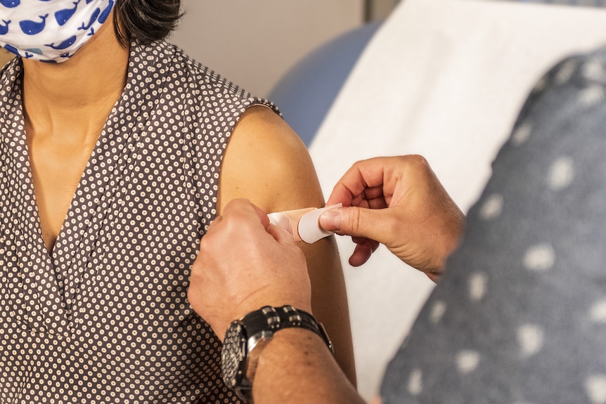 A person applying a bandage on the shoulder of another person who is wearing a mask