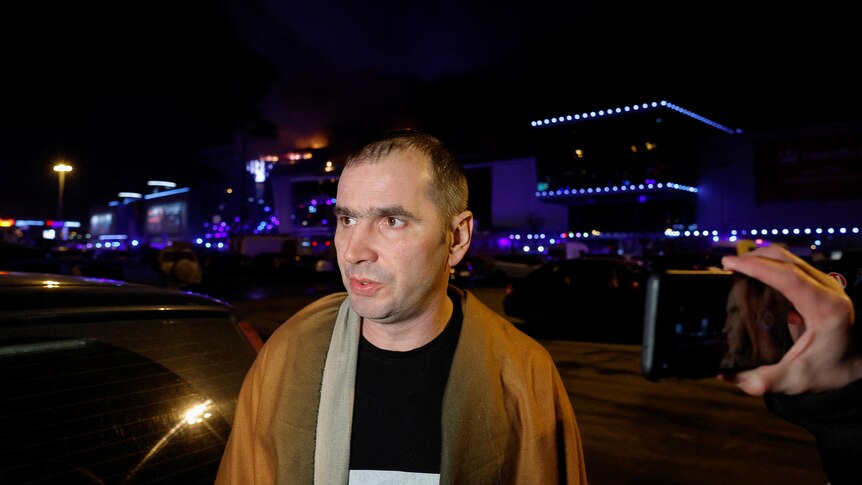 A man stands in a carpark speaking to the media with the glow of a burning building behind him