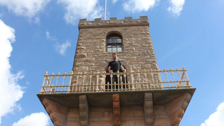 Ben stands on the tower's balcony looking down at the camera and smiling. He is wearing a black shirt and holding the railing.