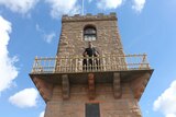 Ben stands on the tower's balcony looking down at the camera and smiling. He is wearing a black shirt and holding the railing.