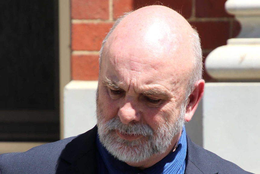 A man in a suit with a grey beard outside a brick building.