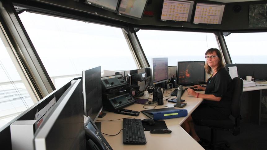 A brown-haired woman with glasses sits t a desk in a control tower for shipping