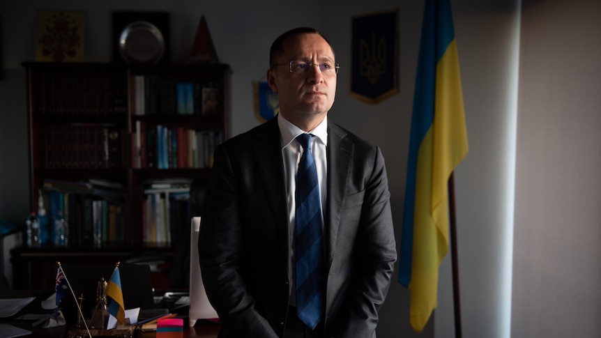 A middle-aged man with short hair and glasses sits on the edge of a desk in front of the Ukrainian flag.