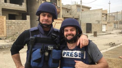 Journalist Matt Brown and cameraman Aaron Hollett stand in an empty street in combat gear front of unfinished buildings.