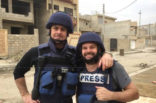 Journalist Matt Brown and cameraman Aaron Hollett stand in an empty street in combat gear front of unfinished buildings.